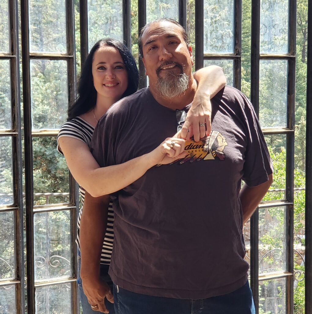 A woman stands behind a man, wrapping her arms around his shoulders. They both smile at the camera in front of large glass windows with a forest view outside, capturing the warmth of Monument Business Alliance in Colorado Springs.