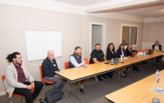 A group of people sit around a rectangular conference table in a meeting room with a whiteboard on the wall.