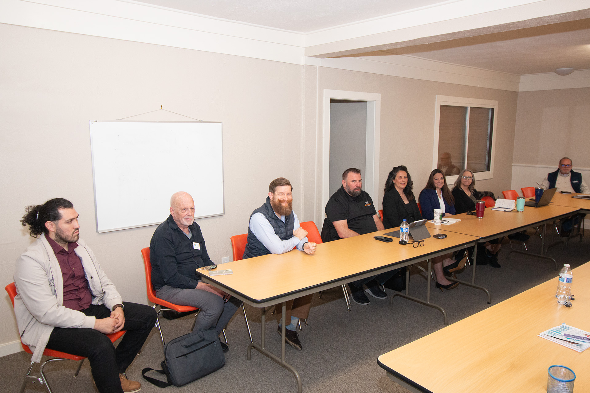 A group of people sit around a rectangular conference table in a meeting room with a whiteboard on the wall.