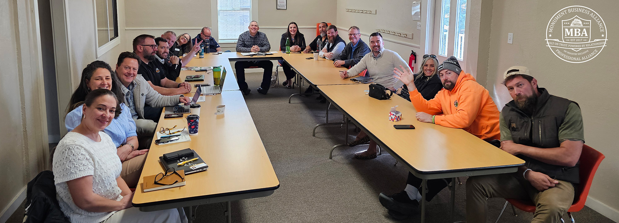 A group of people sit around three tables arranged in a U-shape during a meeting in a classroom or conference room.