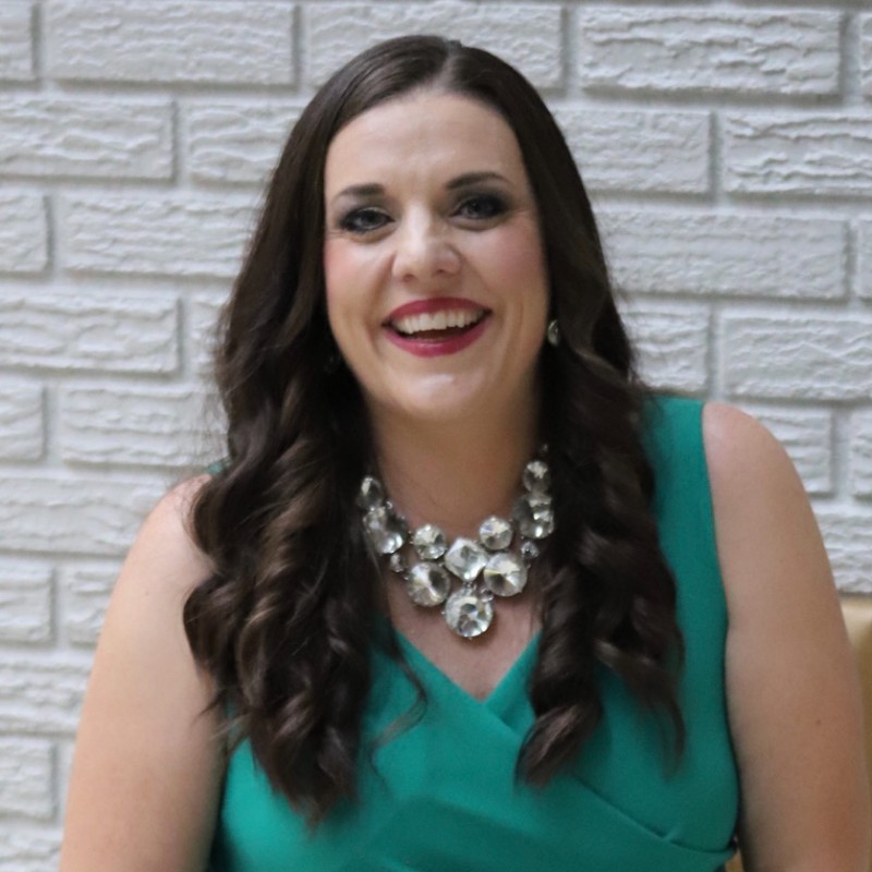 Sarah Fisher Auris Payroll Colorado Springs with long brown hair wearing a green sleeveless dress and a large statement necklace sits in front of a white brick wall, smiling.