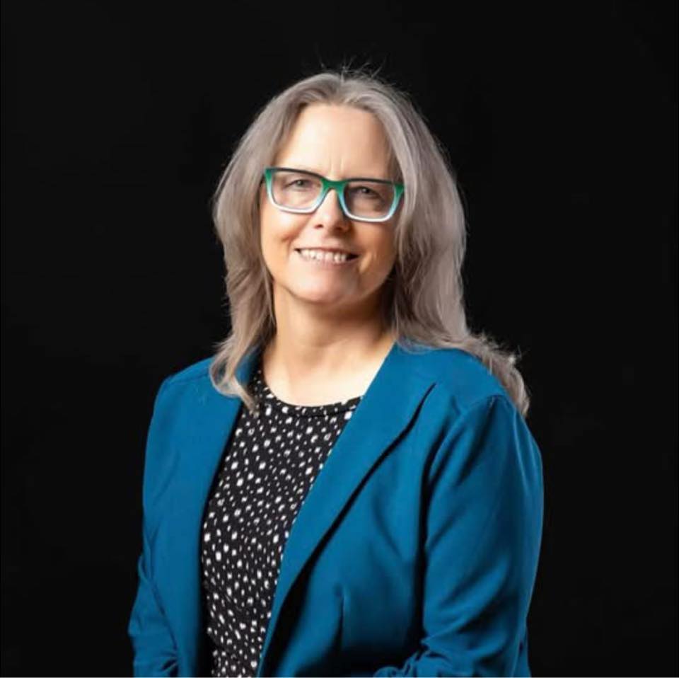 A woman with long gray hair and teal glasses, dressed in a blue blazer over a black and white patterned top, poses confidently against a plain black background—ready for business networking in Colorado Springs.