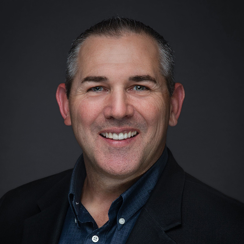 Smiling middle-aged man with short gray hair, wearing a dark blazer and blue collared shirt, posed against a dark background—ideal for Colorado Springs professionals or Monument Business Alliance business networking.