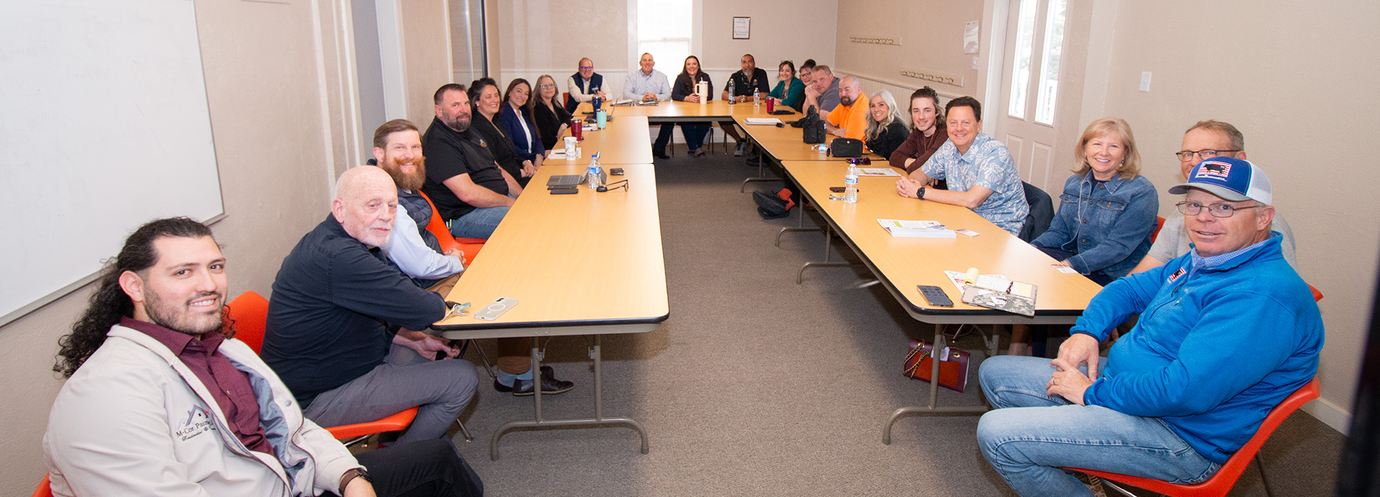 A group of people sit around a large U-shaped table in a meeting room, facing the camera.