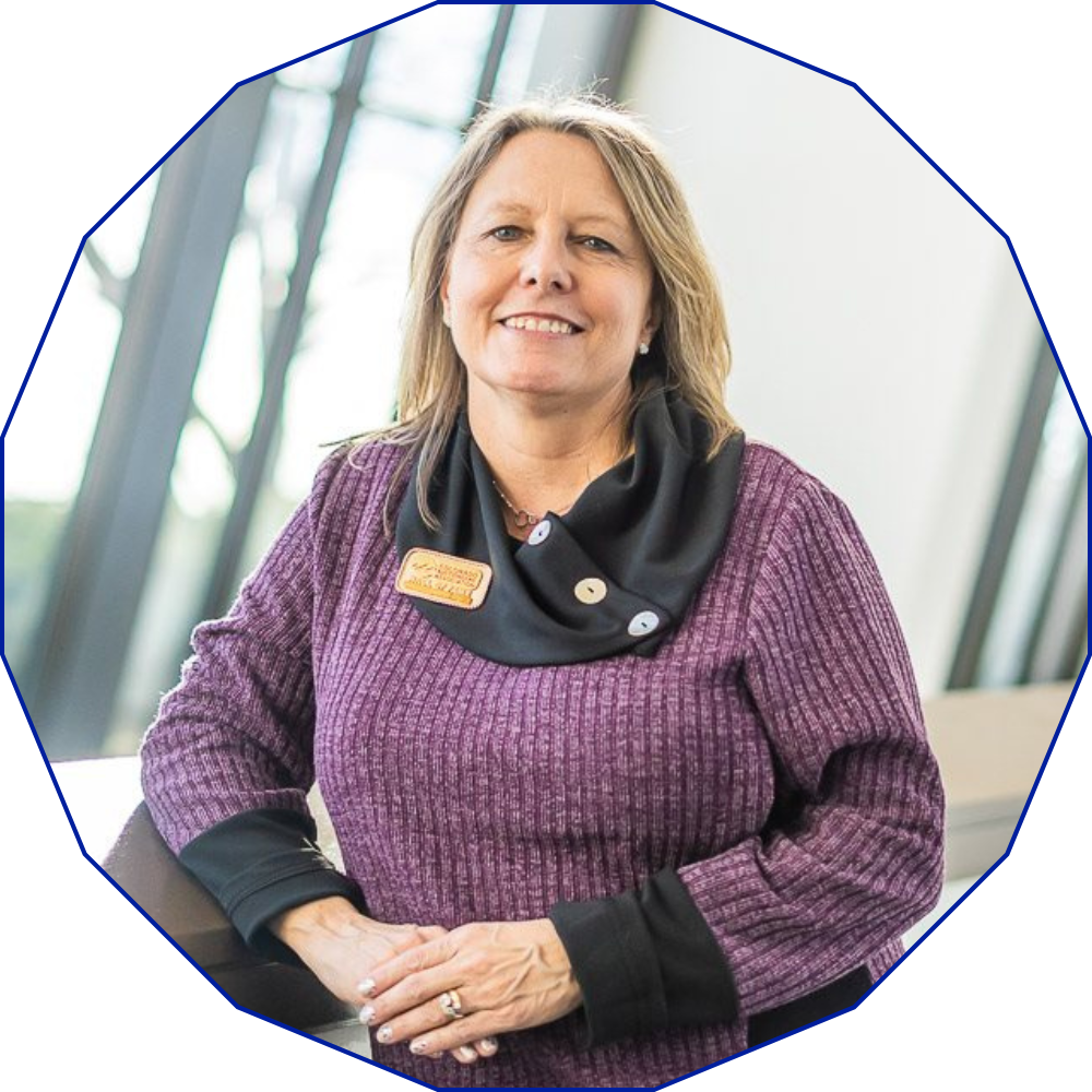 A woman with long blonde hair wearing a purple sweater and black scarf stands indoors, smiling at the camera.