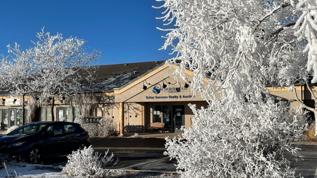 A beige commercial building with signs, a parked black car, and frosted trees in front on a clear, sunny winter day.