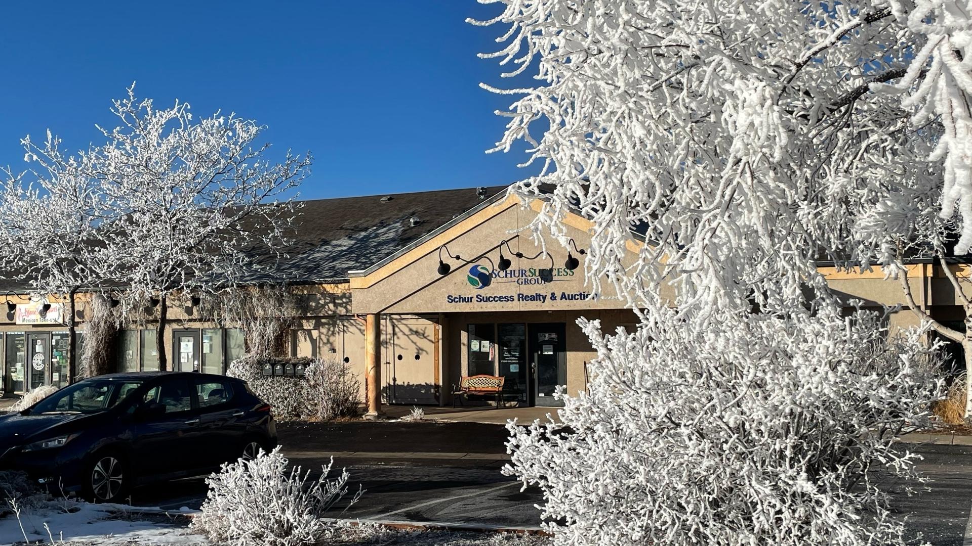 A beige commercial building with signs, a parked black car, and frosted trees in front on a clear, sunny winter day.
