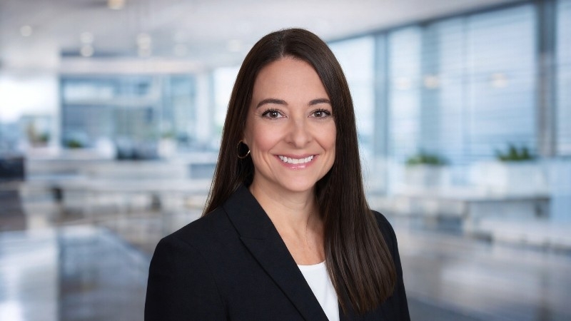 A woman with long brown hair, wearing a black blazer and white top, smiles at the camera in a bright, modern office setting.