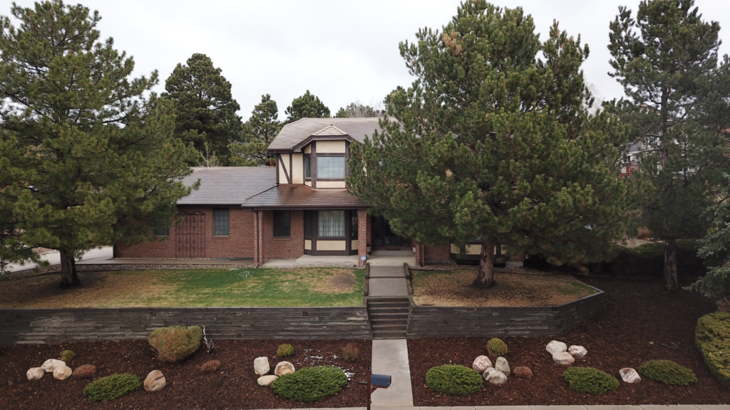 Two-story brick and wood house with a front lawn, large trees, a stone walkway, and landscaped yard on a cloudy day.