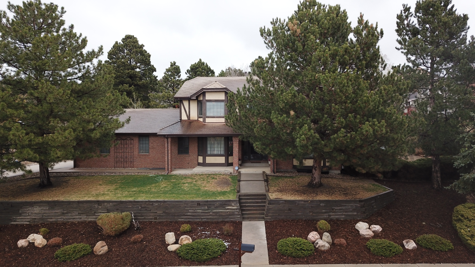 Two-story brick and wood house with a front lawn, large trees, a stone walkway, and landscaped yard on a cloudy day.