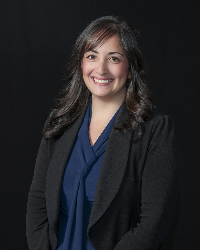 A woman with long dark hair wearing a dark blazer and blue blouse smiles at the camera against a plain dark background.