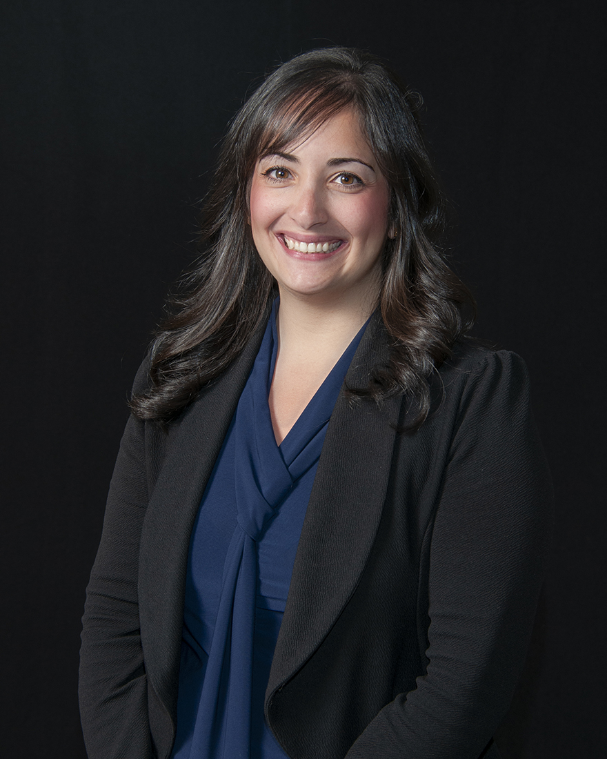 A woman with long dark hair wearing a dark blazer and blue blouse smiles at the camera against a plain dark background.