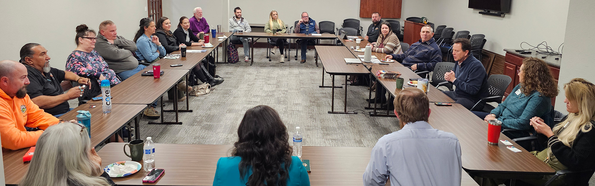 A group of people sit around a U-shaped arrangement of tables in a meeting room, listening to a man in a suit who is speaking.