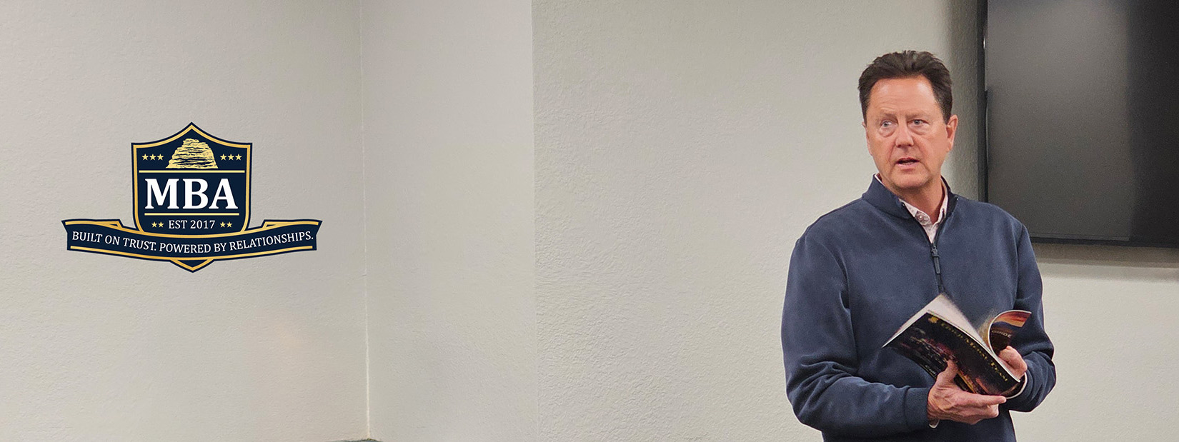 A man in a blue jacket holds an open book and speaks in a classroom; an MBA program logo is displayed on the wall.