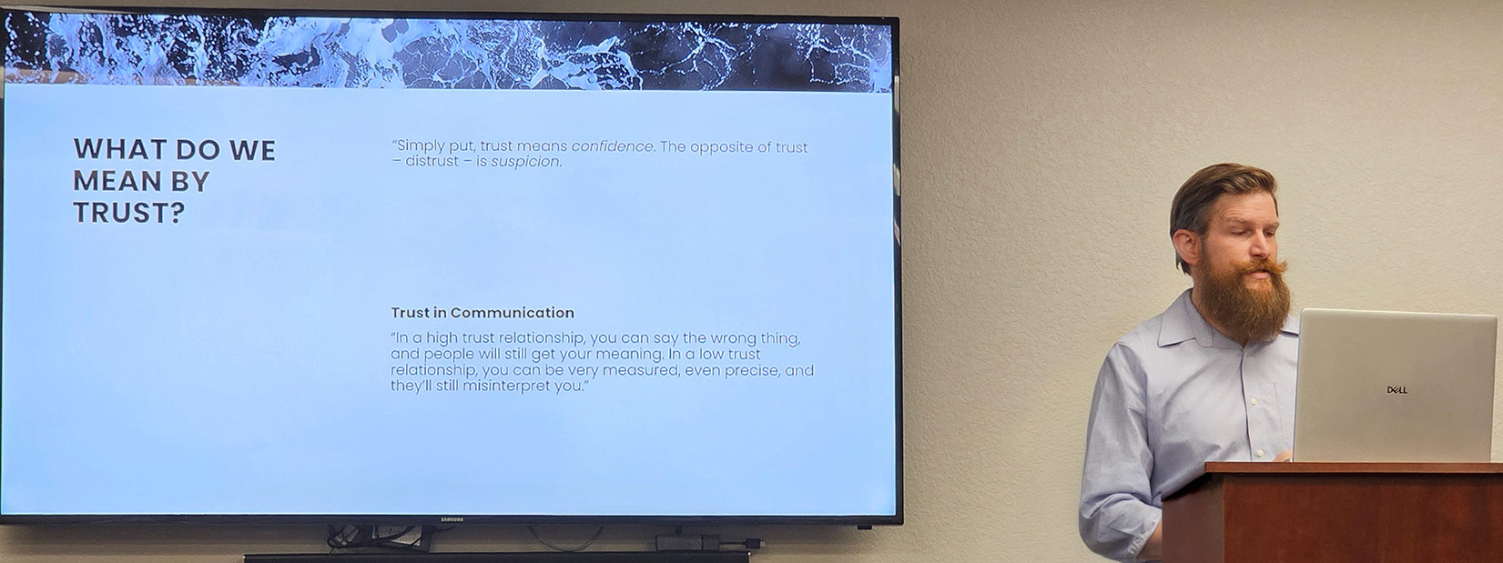 A man with a beard stands at a podium presenting a slideshow titled "What do we mean by trust?" on a large screen in a conference room.