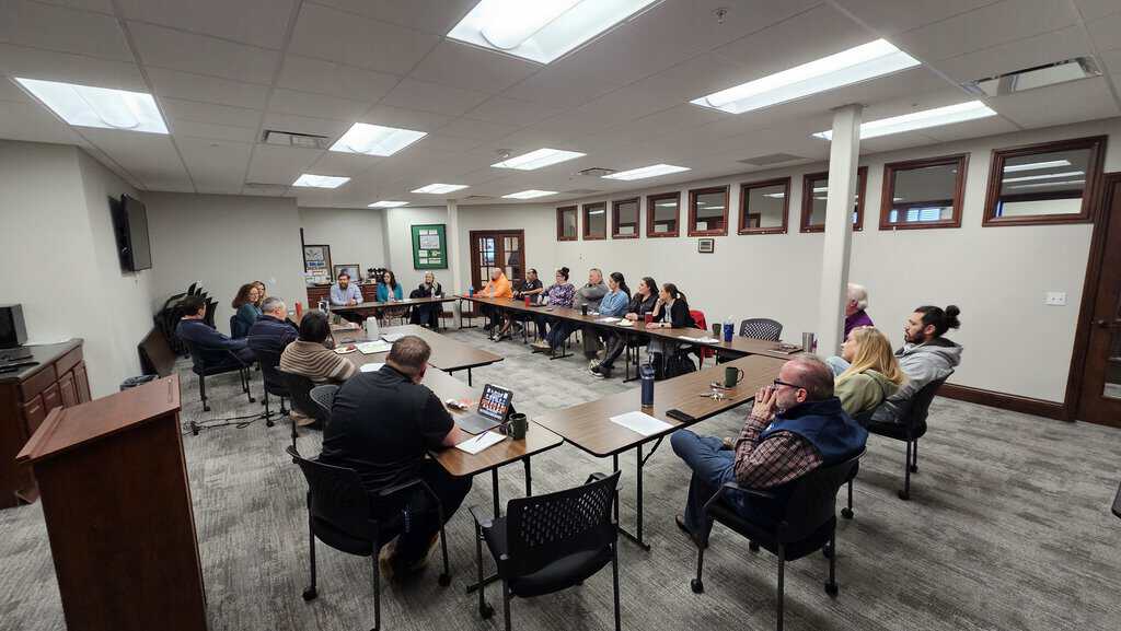 A group of people sit around a U-shaped table in a conference room, engaged in a meeting or discussion.