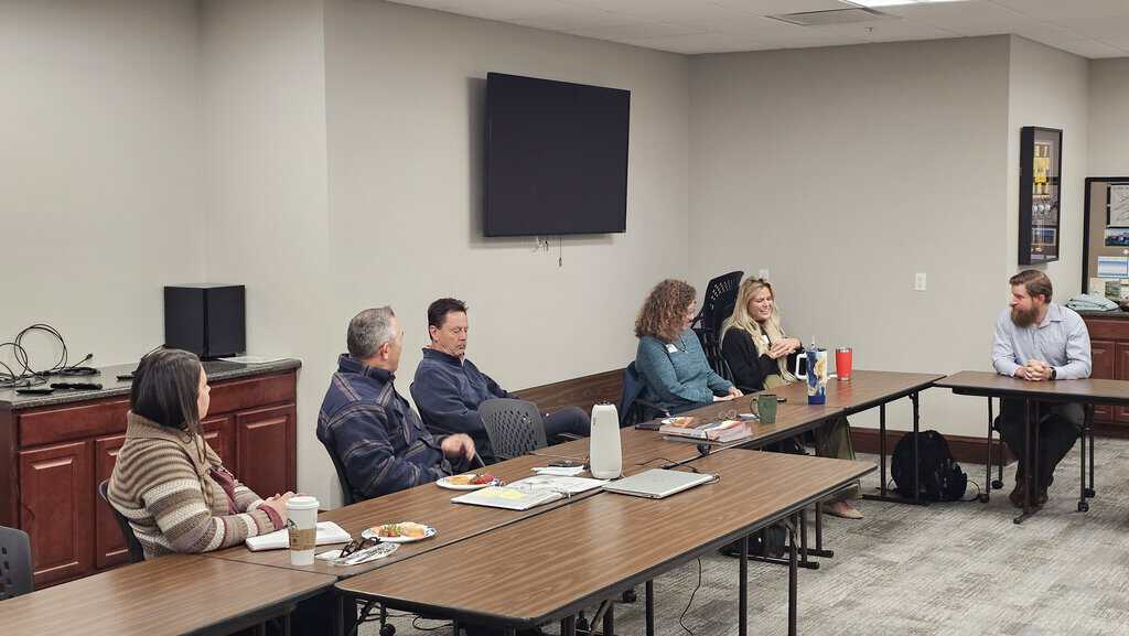 Six people sit around a conference table in a meeting room, engaged in discussion. There are drinks, plates, and papers on the table, and a blank TV screen on the wall.