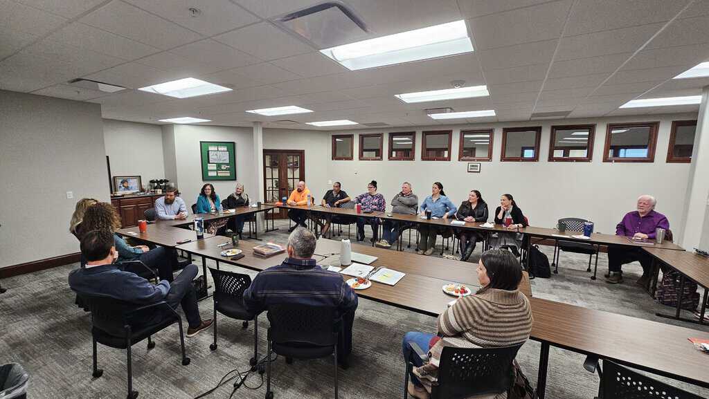 A group of people sit around a large U-shaped table in a conference room, engaged in a meeting or discussion.